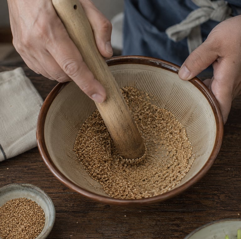 Chef using a wooden pestle to grind toasted sesame seeds in a ceramic suribachi mortar bowl.