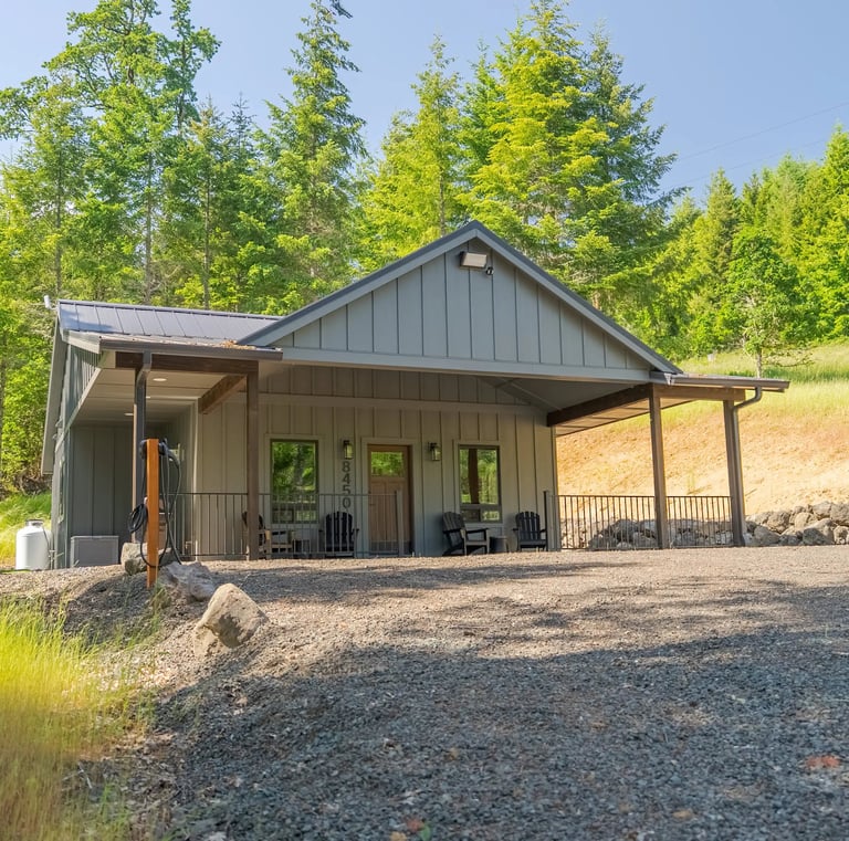 A modern gray board and batten cabin with a metal roof nestled in a lush green forest clearing.