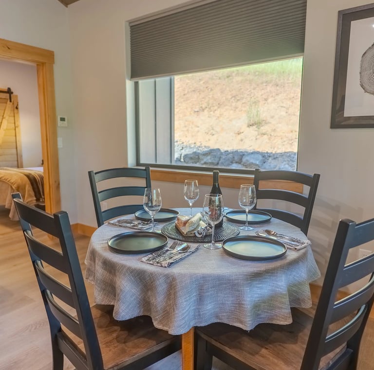 Modern rustic dining area with a round table, black chairs, and place settings near a window.