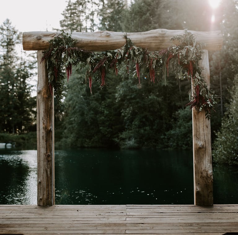 Rustic wooden wedding arch with floral greenery on a lake dock surrounded by trees.