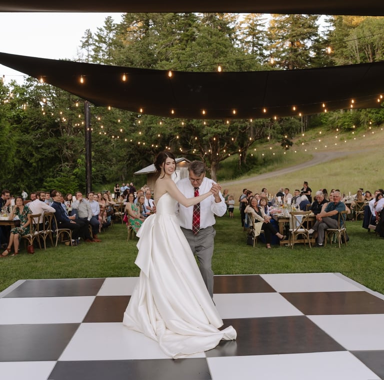 Bride and father dancing on a checkered dance floor at an outdoor wedding reception with string lights.
