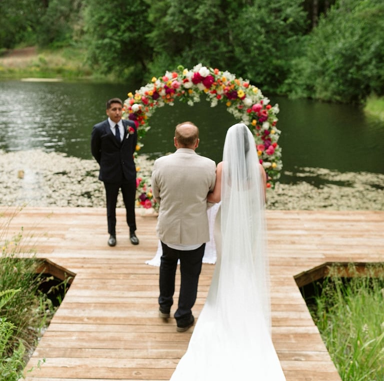 Bride in a white gown walks down a wooden pier toward a floral wedding arch by a scenic lake.