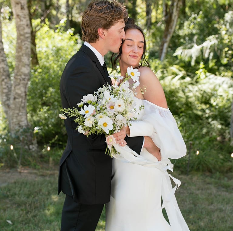 Groom in black tuxedo kissing bride in off-the-shoulder white wedding dress holding a daisy bouquet.
