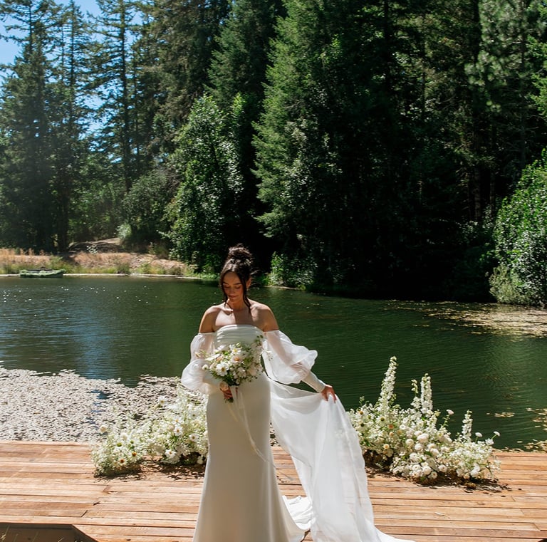 Bride in a white off-the-shoulder wedding dress posing on a wooden dock by a forest lake.