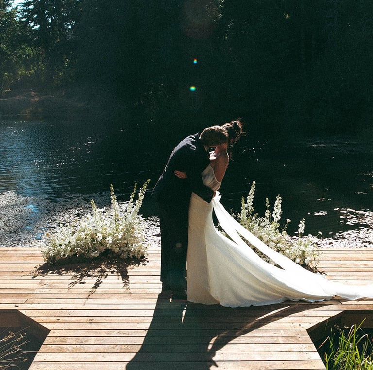A bride and groom kiss on a wooden dock by a lake during an outdoor wedding ceremony.