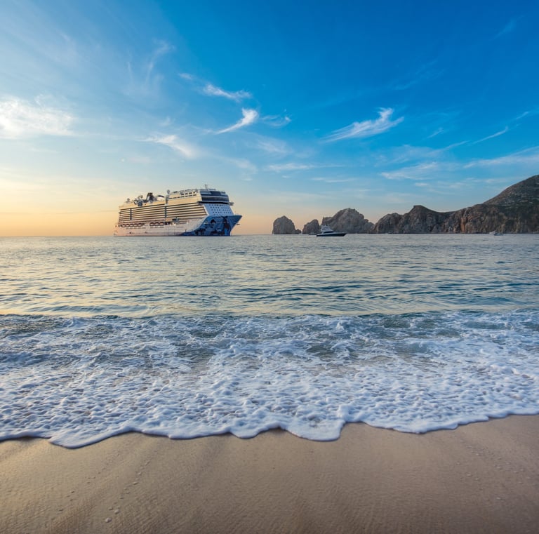 A large luxury cruise ship anchored in the ocean near a tropical beach and rocky cliffs at sunset.