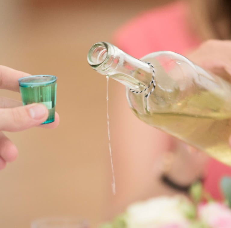 A hand pouring a clear alcoholic beverage from a glass bottle into a small blue shot glass.