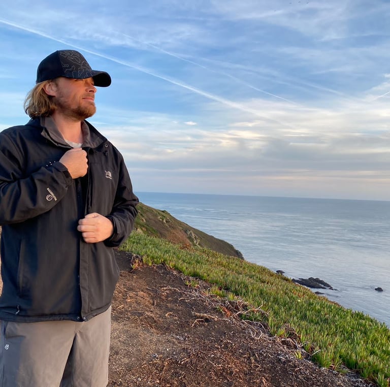 A man in a hiking jacket and cap stands on a coastal cliff overlooking the ocean at sunset.