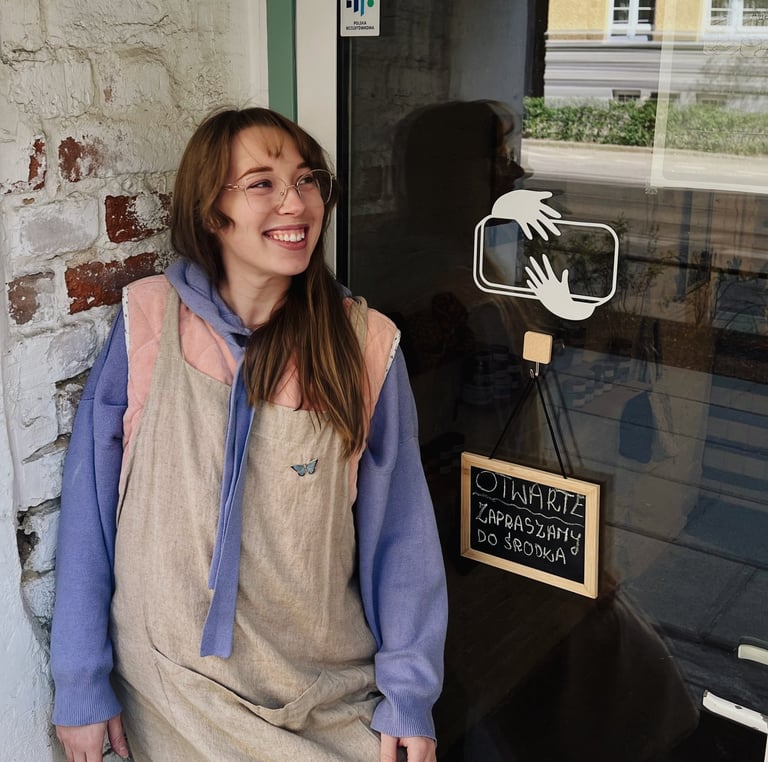 Smiling woman in glasses standing by a shop door with an open sign in Polish.