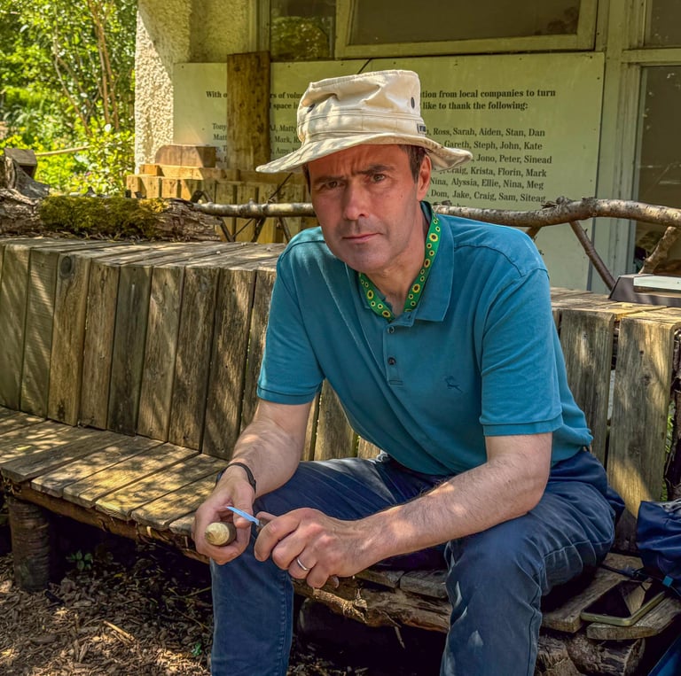 Simon is sitting on a wooden bench in the shade at Westbury Wildlife Park, whittling a mushroom.