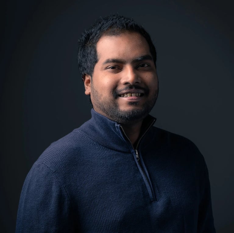 Professional headshot of Tharinda H.A smiling in a navy sweater against a dark gray background.