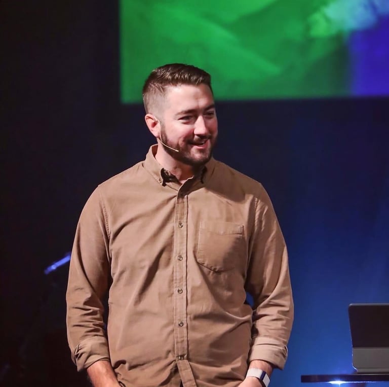 Cole speaking on stage at a ministry event, wearing a tan shirt with a lapel microphone.
