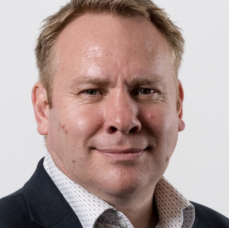 Professional headshot of a middle-aged man with short blonde hair wearing a suit jacket and collared shirt.
