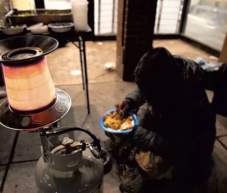 Man sitting on the ground eating his plate of food in front of a propane heater.