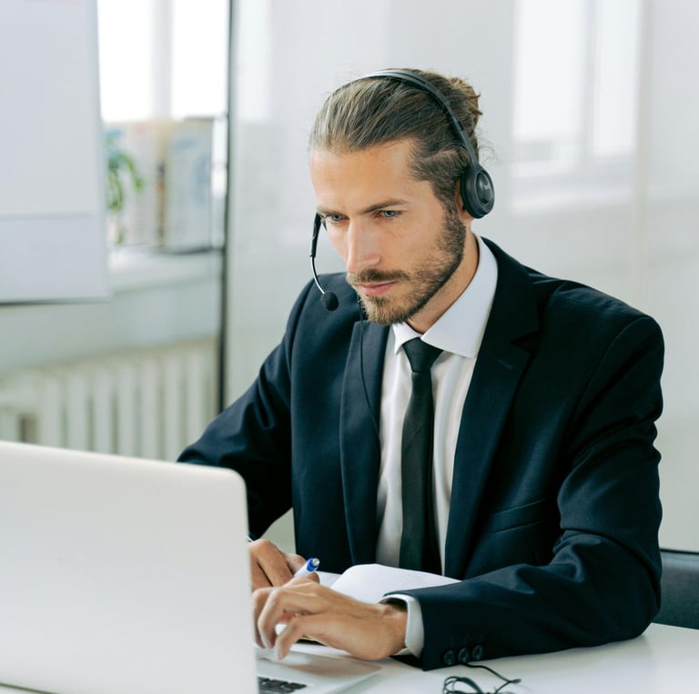 A man with a beard and long hair tied back is wearing a suit and headset while working on a laptop. He holds a pen and appear