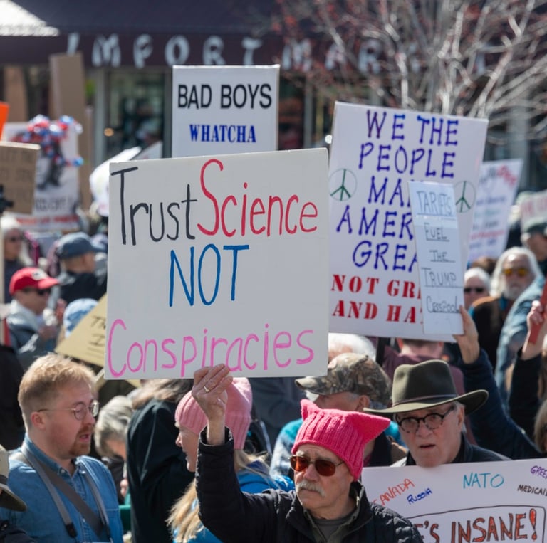 Protesters at a political rally holding a Trust Science Not Conspiracies sign among a diverse crowd.