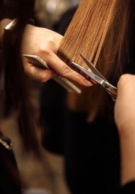 Estilista profesional recortando cabello largo castaño con tijeras de salón y un peine.