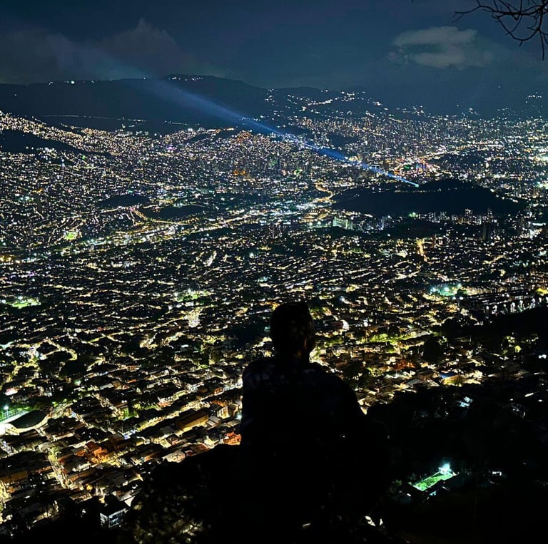 Silhouette of a person overlooking the illuminated city skyline of Medellin at night from a mountain viewpoint.