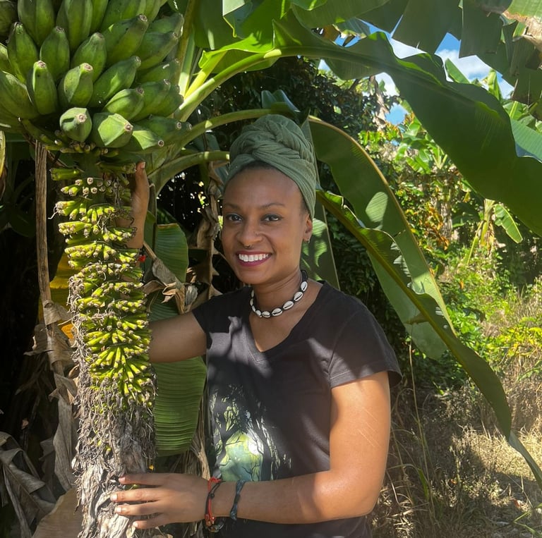 Evvie, a brown-skinned black femme person in a green head-wrap stands underneath a banana tree
