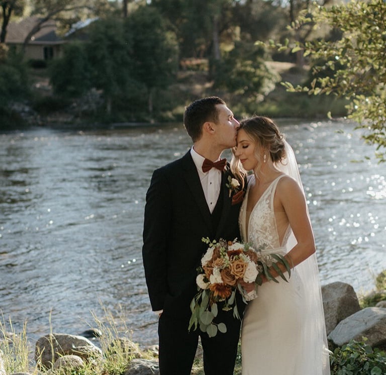 Groom kisses bride on the forehead during a scenic outdoor riverside wedding ceremony.