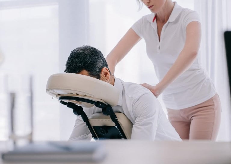 Professional massage therapist performing a seated chair massage for a businessman in an office.