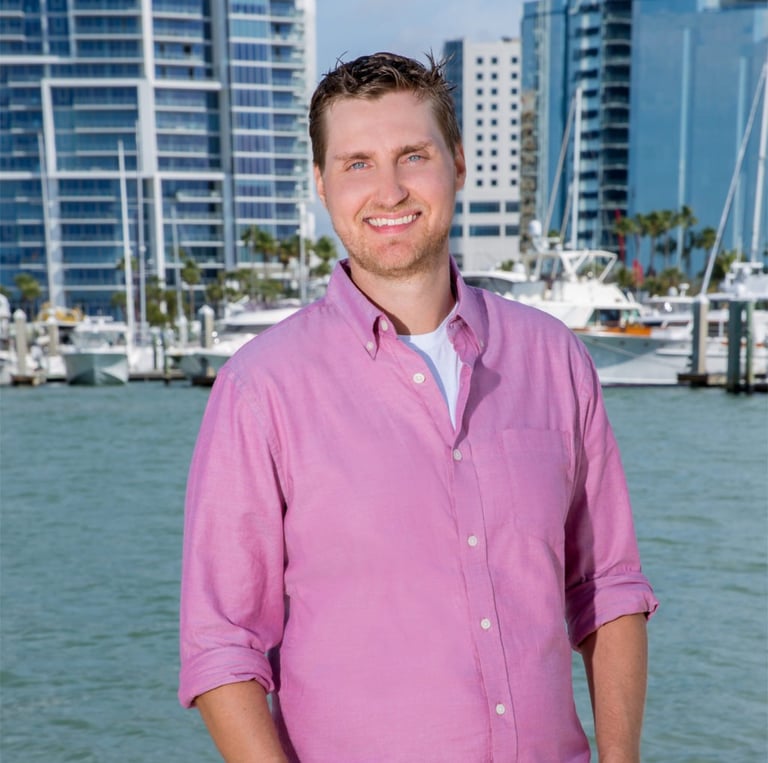 Josiah Ryan, business consultant wearing a pink shirt and jeans standing in front of a body of water