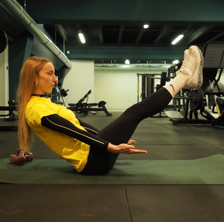 Una donna in maglia gialla esegue l'esercizio 'boat pose' su un tappetino da yoga in palestra, concentrandosi sull'equilibrio