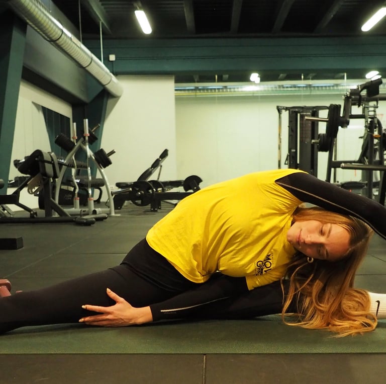 Una donna su un tappetino da yoga in palestra esegue una posa laterale di stretching, tenendo il piede con una mano mentre si