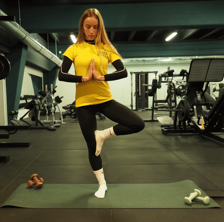 Una donna in posa di yoga albero (Vrksasana) su un tappetino da yoga in palestra. Indossa una maglietta gialla con logo 'Move