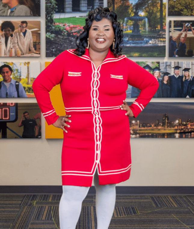 A smiling woman in a stylish red knit dress stands before a wall of campus photography.