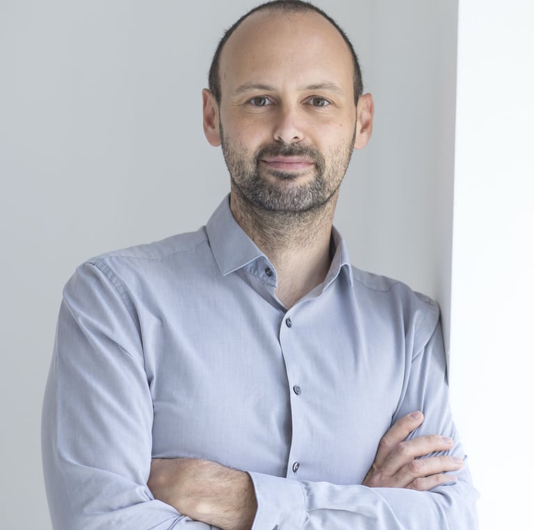 Professional headshot of a smiling man with a beard wearing a grey button-down shirt with arms crossed.