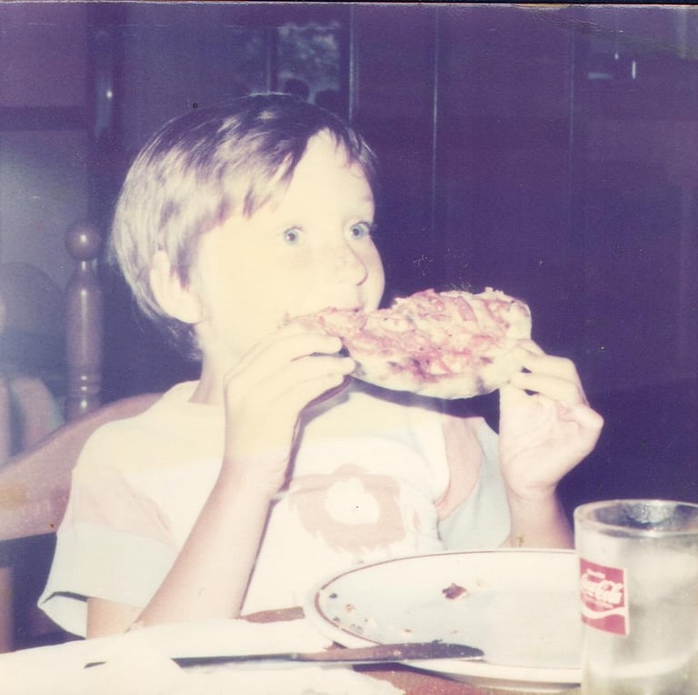 Vintage photo of a young boy eating a slice of pizza at a dinner table.