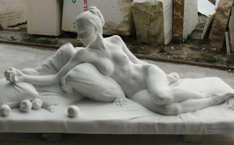 A detailed white marble sculpture of a reclining nude woman resting on pillows in a stone workshop.