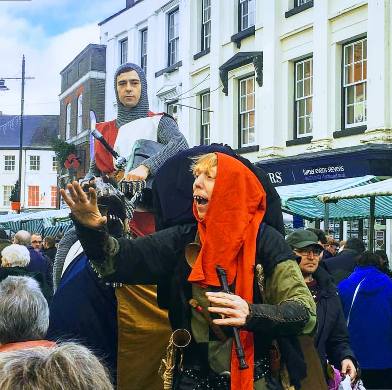 Medieval reenactors in costume including a knight and jester at a crowded outdoor heritage street market.