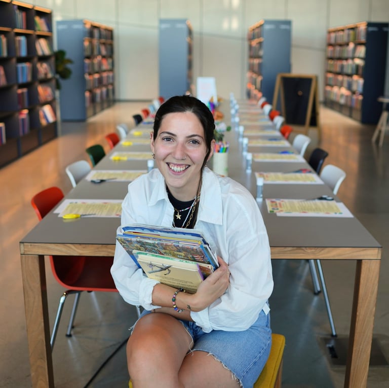 Smiling woman holding a stack of books while sitting in a modern library with study tables.