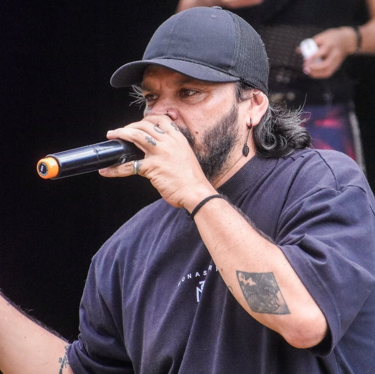 Vocalista masculino con barba, gorra negra y camiseta negra interpretando una presentación en vivo 