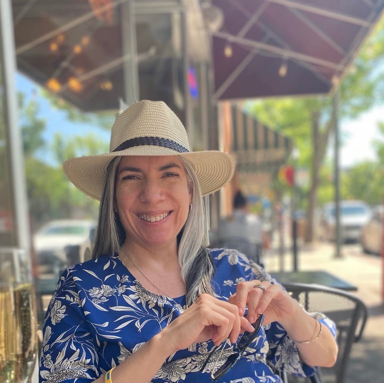 Poet Sheena Powell smiling at an outdoor cafe, wearing a blue patterned shirt and a straw hat