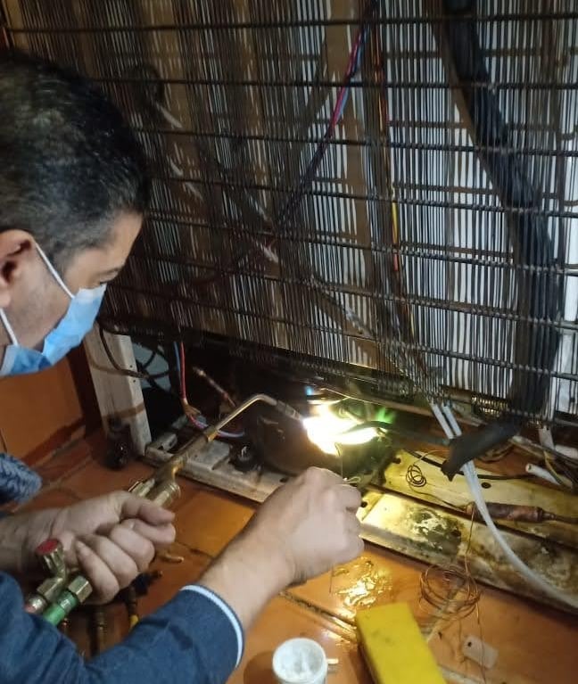 A technician wearing a mask uses a welding torch to repair a refrigerator compressor and cooling coils.