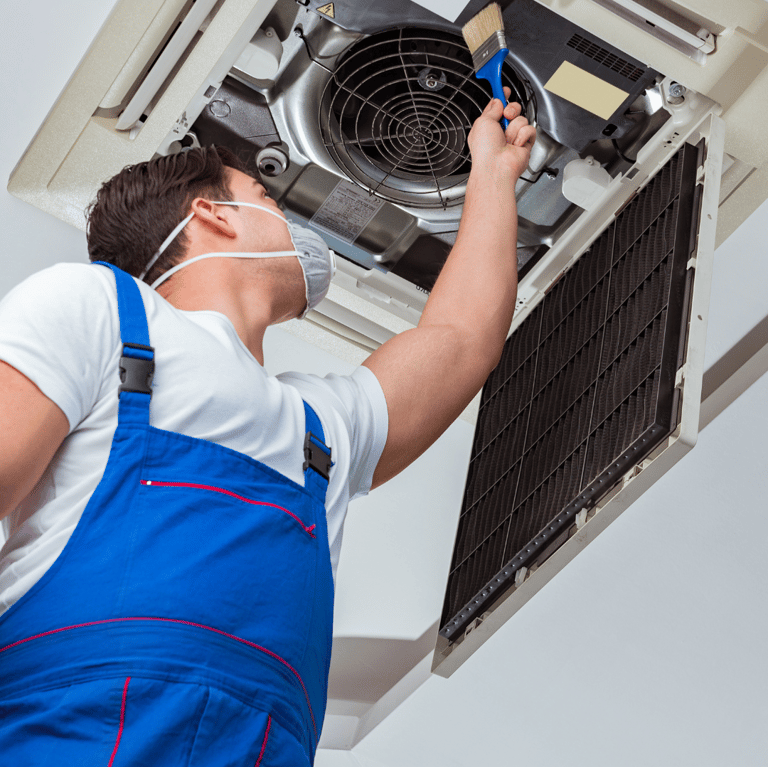 Un hombre con un mono azul y una máscara limpiando equipo de aire acondicionado tipo cassete