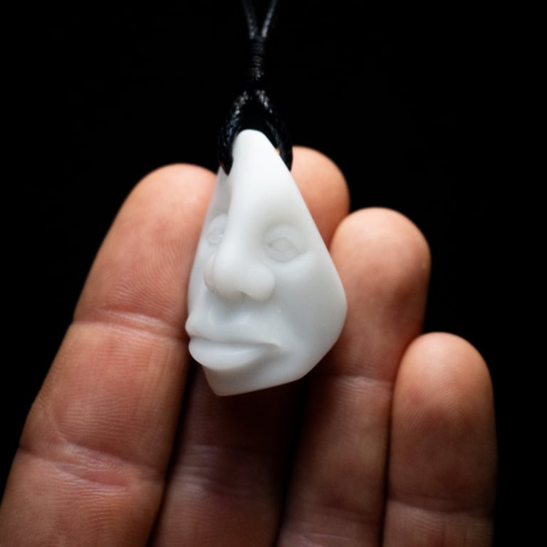 A hand holding a white hand-carved face pendant made of stone on a black cord necklace.