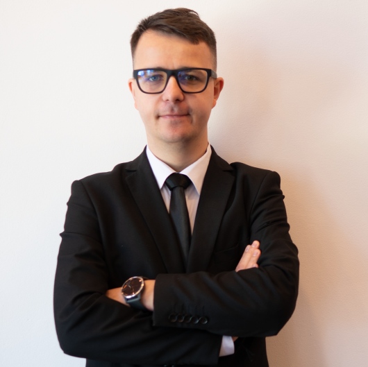 Professional businessman in a black suit and glasses posing with arms crossed against a white wall.
