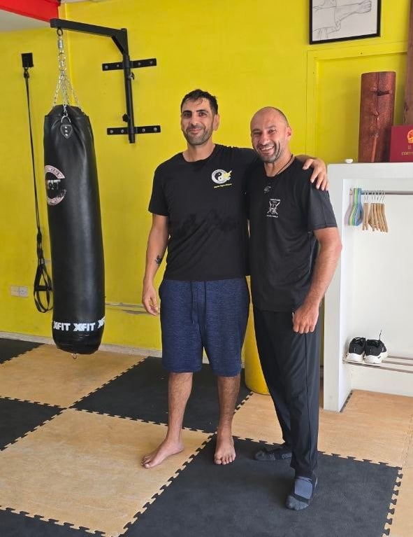 Two smiling martial arts students standing next to a heavy punching bag in a bright training gym.