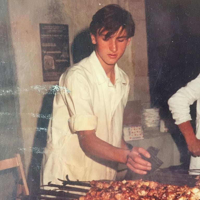 A vintage photo of a young chef in a white uniform grilling meat skewers on an open flame.