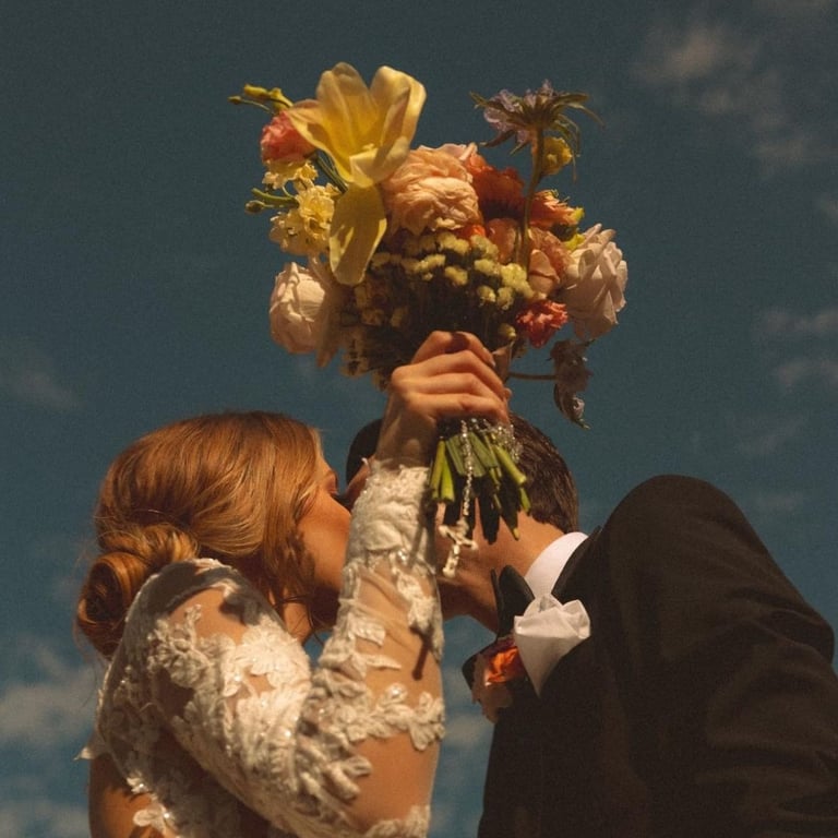 Novia levantando su ramo mientras besa al novio bajo el cielo azul.