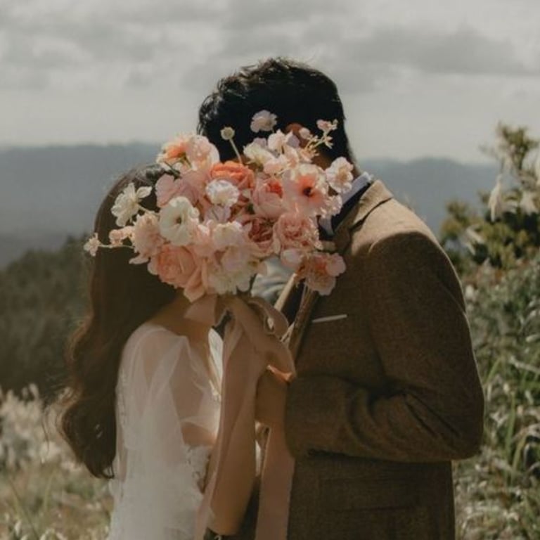 Novios besándose con ramo de flores rosadas frente a paisaje natural.