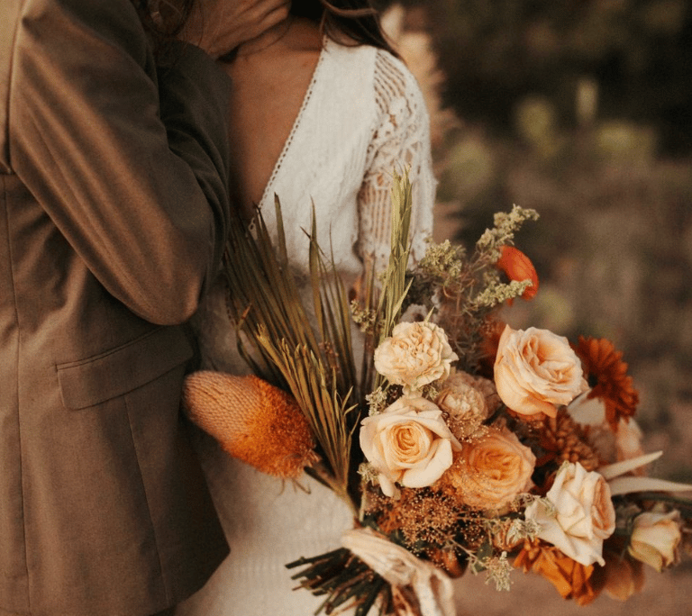 Pareja abrazada con ramo en tonos terracota y crema al atardecer.