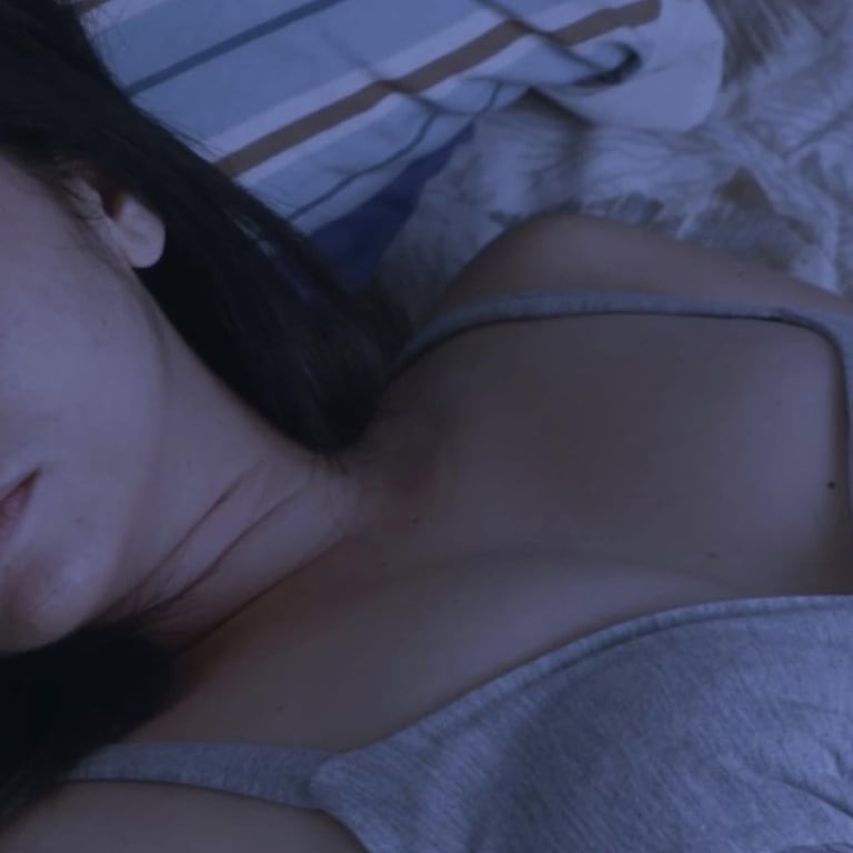 A dark-haired woman wearing a thin grey tank top is asleep in bed
