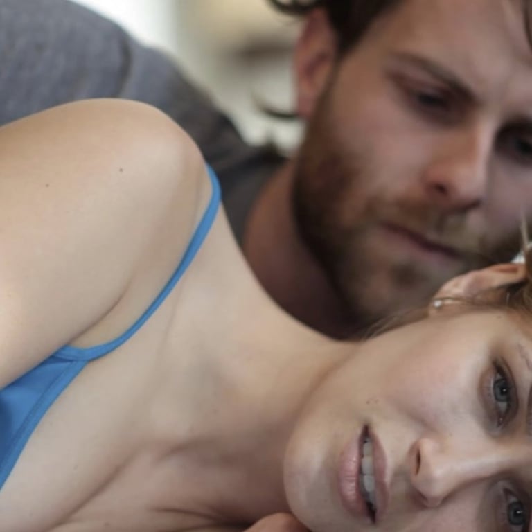 A bearded 30-something man lies on a bed looking down on the 30-something woman lying beside him. Neither one looks happy.