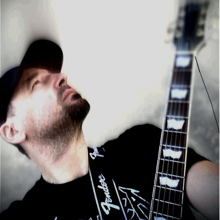 A man wearing a black baseball cap and t-shirt playing an electric guitar with a Fender strap.