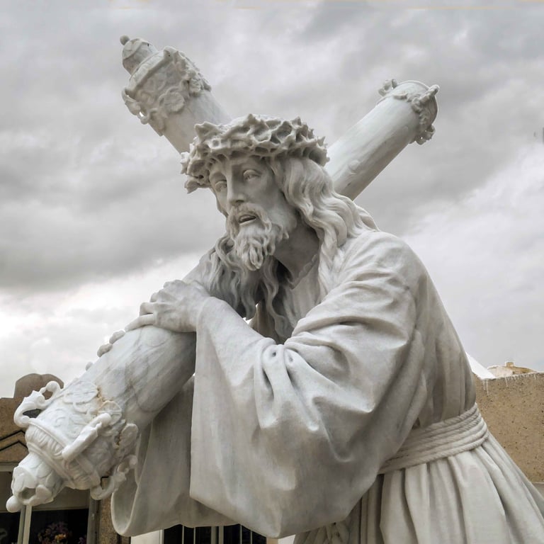 White marble statue of Jesus Christ carrying the cross with a crown of thorns against a cloudy sky.